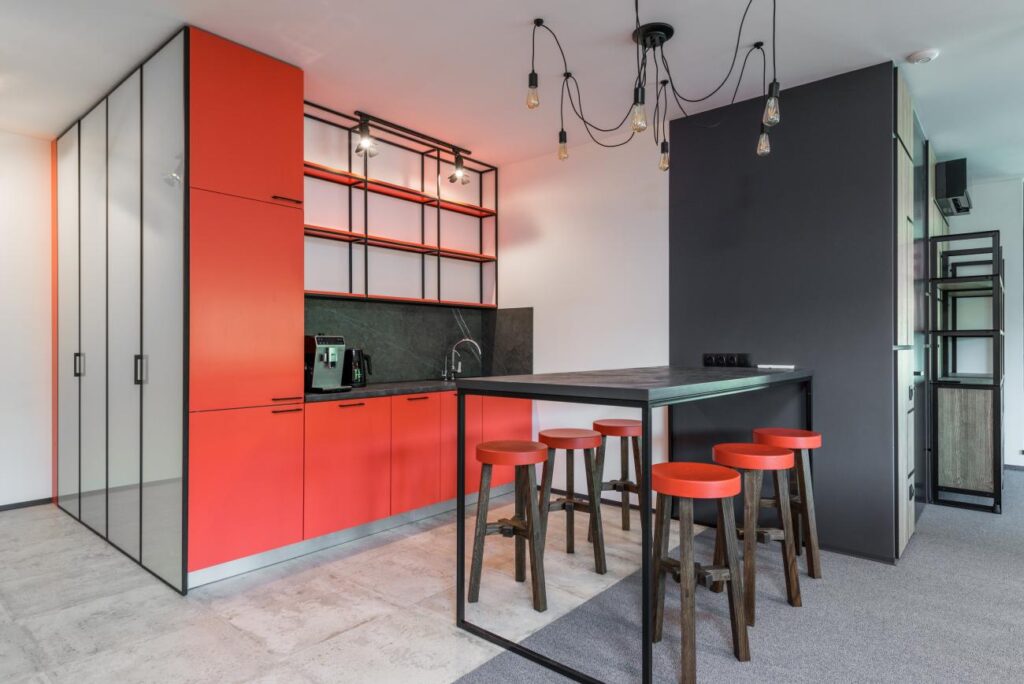 A modern kitchen featuring eye-catching red and gray cabinets, a central island with a black countertop, red stools, and an industrial-style lighting fixture above. 