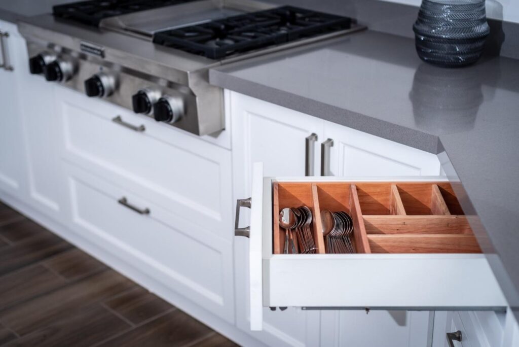  A close-up view of a kitchen's white cabinet with a hidden drawer pulled out, revealing neatly organized silverware in a wooden organizer, adjacent to a stainless-steel gas range.