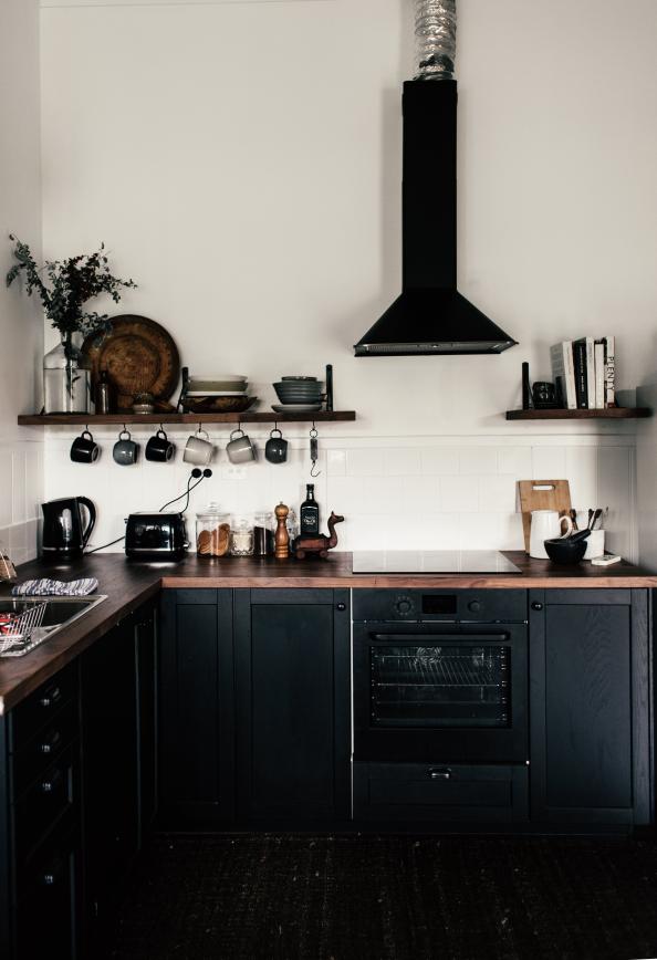 A cozy kitchen corner with traditional black cabinets topped by a wooden countertop, complemented by open shelving displaying a variety of kitchenware, and a modern black range hood above. 