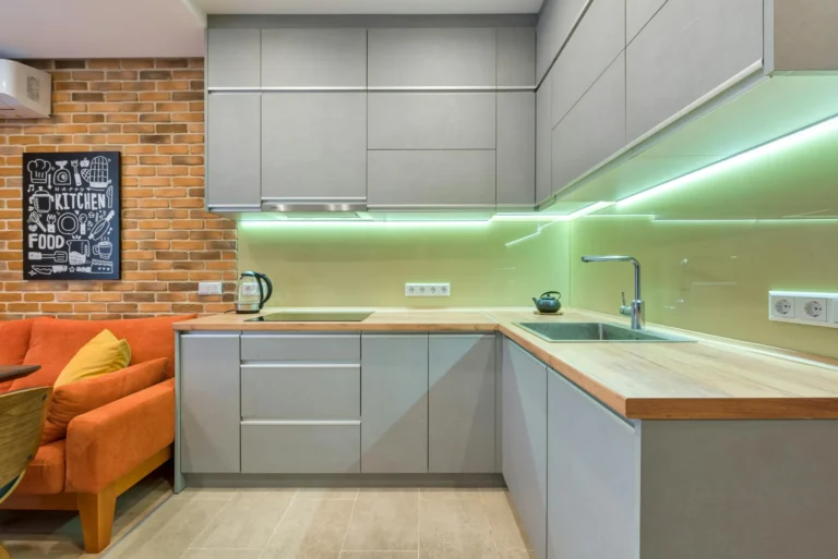 A close-up photo of a modern kitchen with white flat-panel cabinets, stainless steel hardware, and light quartz countertops. A breakfast bar with two modern pendant lights hangs above two wood and chrome bar stools.